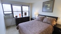This single-floor space showcases a thoughtfully arranged bedroom that balances comfort and functionality. The room is anchored by a queen-sized bed dressed in a warm paisley bedspread, complemented by a dark-toned upholstered headboard and coordinating nightstand. A classic table lamp on the nightstand provides soft ambient lighting, adding warmth to the neutral color palette of white walls and light carpeted flooring.

A prominent double window along one wall floods the room with natural light while framing elevated urban views of the surrounding cityscape, enhancing the sense of openness. Horizontal blinds offer privacy and light control. Beneath the window sits a dark wood multi-drawer dresser, styled with decorative accents including a vase and small figurines, adding a personal touch to the space.

A framed artwork on the adjacent wall contributes visual interest and a polished, finished feel to the room. The overall ambiance is clean, calm, and livable, with dark wood furniture pieces providing a cohesive contrast against the light walls and flooring throughout.