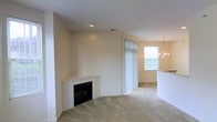 This single-floor residence offers a welcoming and functional layout designed for comfortable everyday living. The main living area is finished with plush wall-to-wall carpeting and centers around a classic tile-surround fireplace, providing a warm and inviting focal point to the room. Neutral, light-toned walls throughout lend the space a clean and versatile aesthetic that adapts easily to a variety of décor styles. Vertical blinds on the sliding door or large window allow for flexible light control while connecting the interior to the outdoors. Recessed lighting in the ceiling provides practical illumination across the living space. The layout flows openly into an adjoining dining area, which is distinguished by a decorative chandelier that adds a touch of elegance. A bar-height counter or partial wall delineates this dining space from what appears to be an adjacent kitchen, promoting a connected yet organized floor plan. Natural light filters in through the dining area window, brightening the space. The consistent carpeting and cohesive color palette unify the living and dining zones seamlessly. Overall, this single-floor home presents a straightforward, well-maintained layout suited for a range of lifestyles.