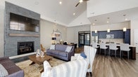 This thoughtfully designed single-floor community space blends coastal charm with modern amenities, offering residents a welcoming and versatile gathering hub. The main lounge is a standout area, featuring soaring vaulted ceilings, a ceiling fan, and a striking floor-to-ceiling stacked stone fireplace topped with a wall-mounted television—creating a warm, inviting focal point. Comfortable seating arrangements, including plush sofas and striped accent chairs, encourage relaxed conversation throughout the space. Adjacent to the lounge, a bright and airy dining area benefits from abundant natural light through large windows, with clean hardwood-style flooring that continues the space's contemporary aesthetic. The full kitchen is a functional showpiece, boasting bold navy cabinetry, a herringbone tile backsplash, stainless steel appliances, and a generous white quartz island with pendant lighting overhead. A quieter lounge area with plush carpeting provides a more intimate setting for small gatherings or relaxation. The fitness center is expansive and well-equipped, featuring treadmills, ellipticals, weight benches, a full dumbbell rack, strength machines, and large mirrors along one wall—all accessible via stylish black-framed glass doors. Natural light filters throughout, enhancing the open, airy feel of the entire facility.