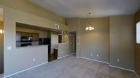 This single-floor space showcases an open-concept design that seamlessly integrates the kitchen and living areas. The living space is finished with neutral-toned carpet, creating a warm and inviting atmosphere, while warm beige walls run throughout, contributing to a cohesive aesthetic. A decorative chandelier with warm-toned bulbs serves as a focal point, adding a touch of elegance to the dining or living area.

The kitchen is positioned adjacent to the main living space, separated by a raised countertop bar that doubles as a partition and casual dining surface. Dark wood cabinetry and a black refrigerator provide contrast against the lighter countertops, while track lighting illuminates the kitchen workspace. A visible HVAC vent is integrated into the upper portion of the kitchen area, ensuring climate control throughout the space.

A hallway visible beyond the kitchen area leads to additional rooms, with track lighting guiding the way. The overall layout is practical and functional, with clear sightlines across the open floor plan, making the space feel connected and well-organized.