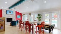 This lively community clubhouse is designed with a bold, resort-inspired aesthetic that immediately draws the eye. The open-plan layout is anchored by striking teal exposed ceiling beams that run the length of the space, complemented by vaulted ceilings fitted with ceiling fans and recessed lighting that keep the atmosphere bright and airy. Vivid red accent walls provide a dynamic contrast against the crisp white walls and light-toned flooring throughout.

The main gathering area offers multiple seating zones, including a set of white high-top tables surrounded by red metal bar stools, ideal for socializing or casual dining. A lounge area features plush teal velvet armchairs arranged around a round wooden coffee table, all positioned beside a full-height stacked stone fireplace with a wall-mounted television above — a perfect spot for relaxation.

A refreshment station includes teal cabinetry, a beverage refrigerator, a coffee maker, and a large refrigerator, all set against a bold red wall with pendant lighting. A vending area with a Pepsi cooler and snack display rounds out the amenities.

Multiple sets of French doors line the far wall, flooding the interior with natural light and offering direct views and access to an outdoor pool and patio area, seamlessly blending indoor comfort with outdoor living.