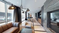 This elegant single-floor space is designed with a contemporary aesthetic, blending refined materials and thoughtful layout to create a versatile lounge and gathering environment. The elongated floor plan is anchored by striking marble tile flooring with dark inlay detailing, flowing throughout the main areas and kitchen zone.

The primary lounge area features a rich brown leather sofa paired with cream and navy accent chairs, illuminated by dramatic multi-arm pendant lighting with industrial-inspired black metal shades. Floor-to-ceiling windows flood the space with natural light and offer views of an outdoor terrace, complete with greenery and a pool beyond.

A secondary seating zone transitions into a dining area, furnished with a mix of teal, navy, and cream upholstered chairs around a central table, set against a backdrop of deep charcoal drapery and a textured stone accent wall. Steel-framed glass partitions add architectural interest while maintaining visual openness.

The fully equipped kitchen features warm wood cabinetry, matte black countertops, a gas cooktop, stainless steel range hood, double wall ovens, and a large island with an undermount sink. Globe pendant lights and open metal shelving complete the refined, functional design. The overall ambiance is polished, modern, and welcoming.