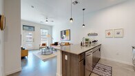 This contemporary single-floor residence is thoughtfully designed with an open-concept layout that seamlessly connects the kitchen, dining, and living areas. The kitchen is a standout feature, showcasing rich dark wood cabinetry extending to the ceiling, stone countertops, stainless steel appliances, and a spacious peninsula with bar seating illuminated by sleek pendant lighting. The living area is bright and welcoming, benefiting from generous natural light streaming through large windows and a glass-panel door that leads to the outdoors. Polished concrete flooring flows consistently throughout the space, lending a clean, industrial-modern aesthetic. The bedroom is a calm retreat, featuring ample natural light from a large window with urban views, neutral tones, and enough space for a full furniture arrangement. A ceiling fan adds comfort to the main living area. The bathroom is finished with large-format tile surrounding a tub-shower combination, a wood-toned vanity with stone countertop, and brushed gold fixtures that add a touch of warmth. Throughout the residence, soaring ceilings amplify the sense of openness, while the cohesive modern design palette of whites, warm woods, and concrete creates a refined, livable atmosphere.
