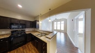 Located at 11289 E Hamilton Crest Blvd in Fishers, Indiana, this single-floor residence offers a thoughtfully designed open-concept layout that seamlessly connects the kitchen and living areas. The kitchen is appointed with rich, dark cabinetry, granite countertops, a double-basin sink, and black appliances, creating a sleek and functional cooking space. A breakfast bar provides a casual dining option and serves as a natural transition point into the main living area. Pendant lighting hangs above the bar, adding warmth and visual interest. A graceful arched passageway frames the transition between the kitchen and the living room, adding an elegant architectural detail to the open floor plan. The living area features vaulted ceilings that lend a sense of spaciousness and airiness to the room, complemented by a ceiling fan for year-round comfort. Large windows along the far wall allow generous natural light to fill the space and offer views of the outdoors. Rich hardwood flooring flows throughout both the kitchen and living areas, tying the spaces together with a cohesive, warm aesthetic. The overall ambiance is inviting and well-appointed, balancing modern finishes with comfortable, livable design.