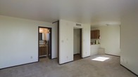 This single-floor unit is laid out to make efficient use of its space, with all key areas accessible from a central open-plan living zone. The floors throughout the main living area are covered in neutral-toned carpet, lending a quiet, comfortable feel underfoot. White walls with dark trim accents define the interior aesthetic, creating a clean and understated look throughout.

Positioned to the right of the main living area, the kitchen is equipped with warm wood-toned cabinetry that provides ample storage space. A countertop with workspace separates the kitchen from the living area, contributing to an open and connected flow between the two zones. A ceiling-mounted HVAC vent is visible, ensuring climate control throughout the space.

A built-in closet with a rod for hanging storage sits centrally along the back wall, offering practical organization directly accessible from the main living area. Adjacent to the closet, a doorway leads to a bathroom that features a vanity with a sink and mirror, warm wood cabinetry below, and tile flooring. Natural light filters into the main living area, adding brightness to the overall atmosphere of the space.