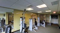 This fitness facility offers a dedicated space for strength training and exercise, thoughtfully designed to accommodate a variety of workout routines. The room features a collection of Hoist-brand strength training machines, including cable systems, chest press, and seated exercise equipment, arranged to allow comfortable movement between stations.

The space is finished with neutral-toned carpeting and warm olive-green accent walls, creating a focused yet inviting atmosphere. A dropped ceiling with recessed panel lighting provides consistent, even illumination throughout the room. A large mirror along one wall enhances the sense of space and allows users to monitor their form during workouts.

Ventilation is integrated into the ceiling, ensuring a comfortable environment during exercise. A glass-paneled door at the far end of the room offers access to an adjacent area, contributing to the overall flow of the facility. Decorative wall art adds a subtle design touch, balancing the functional nature of the space. Overall, the fitness center is well-organized and equipped to meet a range of strength training needs in a clean, professional environment.