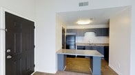 This studio offers a thoughtfully designed single-floor layout that maximizes functionality within a compact yet comfortable footprint. The open-concept floor plan seamlessly connects the living area to a modern kitchen, creating a cohesive and efficient living space.

The kitchen is a standout feature, showcasing sleek gray cabinetry that extends to the upper and lower walls, complemented by a classic white subway tile backsplash that adds texture and visual interest. Black appliances, including a full-size refrigerator and dishwasher, provide a bold contrast against the lighter elements of the space. A peninsula breakfast bar with a granite-style countertop extends into the living area, offering additional prep space and casual seating — ideal for dining or working from home.

Warm hardwood-style flooring flows throughout the kitchen and main living areas, while neutral-toned carpet adds comfort underfoot in the designated living zone. The walls are finished in crisp white, amplifying the sense of openness and allowing natural light to reflect throughout the space. Ceiling-mounted lighting ensures the interior remains bright and welcoming. A textured ceiling and flush-mounted light fixture complete the kitchen's practical yet polished aesthetic, making this studio a functional and inviting place to call home.