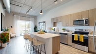 Located at 301 Second St SW in Winter Haven, Florida, this contemporary single-floor residence showcases a thoughtfully designed open-concept layout with impressive soaring ceilings that lend an airy, loft-like feel throughout. The kitchen is a standout feature, offering light wood cabinetry, a white subway tile backsplash, quartz countertops, and a full suite of stainless steel appliances including a range, microwave, and dishwasher. A generously sized central island with bar seating serves as both a functional prep space and a casual dining area, anchoring the open living and kitchen zone. Track lighting and a pendant light above the island add dimension to the industrial-inspired ceiling detail. The living area is bright and welcoming, with large sliding glass doors that invite natural light and provide views of the outdoor surroundings. Wood-look flooring runs throughout, tying the spaces together cohesively. The bedroom is calm and comfortable, featuring tall windows with sheer curtains, a walk-in closet, and soft neutral tones. The bathroom is clean and modern, with white tile, a glass-enclosed shower, and warm wood vanity cabinetry that echoes the kitchen's design language. An in-unit washer and dryer are also present, adding convenience to this well-rounded residence.
