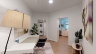 This thoughtfully designed single-floor residence offers a clean, contemporary aesthetic throughout, characterized by crisp white walls, light wood-tone flooring, and an abundance of natural light. The open-concept living area serves as the heart of the home, featuring large sliding glass doors that lead to a private balcony, flooding the space with daylight and providing an inviting indoor-outdoor connection. The kitchen is outfitted with stainless steel appliances — including a full-size refrigerator, gas range, and built-in microwave — complemented by light wood cabinetry that extends to the ceiling for generous storage. The two bedrooms are comfortably proportioned and bathed in natural light from large windows. One bedroom includes a dedicated walk-in closet with ample hanging space. The bathroom features a sleek, minimalist design with an undermount sink set into a clean white countertop and a backlit vanity mirror. A dedicated workspace area provides a functional home office nook. Completing the unit is an in-unit stacked washer and dryer tucked neatly into its own closet alcove, offering maximum convenience. The cohesive design palette and efficient layout make this a highly functional and visually polished living space.