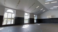 This single-floor multipurpose hall is defined by its generous open layout and impressive vertical scale, with soaring ceilings that amplify the sense of space throughout the interior. The room features a striking two-tone wall treatment, with crisp white upper walls contrasting against a bold black lower panel that runs the length of the space, lending a clean and functional aesthetic. A row of tall arched windows along one wall allows abundant natural light to pour in, casting bright patterns across the floor and creating a well-lit environment during daytime hours. Supplementing the natural light, rows of linear fluorescent fixtures are evenly distributed across the angled ceiling, ensuring consistent illumination throughout. The floor is finished with a smooth, durable surface featuring painted court markings in blue, indicating its use for athletic or recreational activities. The expansive, unobstructed floor plan makes this hall highly versatile, accommodating sports, fitness classes, community gatherings, or large events. The combination of high ceilings, natural light from the arched windows, and ample artificial lighting creates an open, energetic ambiance well-suited to active and dynamic uses.