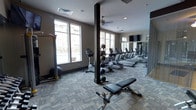 This fitness center is designed to support a comprehensive workout experience within a single, open-plan floor. The space is finished with durable, patterned carpet tiles that add a contemporary aesthetic while providing comfort underfoot. Large windows along one wall flood the room with natural light and offer views of the surrounding exterior, creating an airy, motivating atmosphere. A ceiling fan aids in maintaining comfortable airflow throughout the space.

The cardio section is well-stocked with a row of treadmills and elliptical machines aligned along a full-length mirrored wall, which visually expands the space and allows users to monitor their form. A flat-screen television is mounted high on the mirrored wall for entertainment during workouts. The strength and flexibility area includes a stationary bike, a rowing machine, a stability ball, and an adjustable weight bench, offering a range of training options. Dark structural columns punctuate the space without disrupting the open layout. Recessed lighting complements the natural light, ensuring consistent illumination throughout the room.