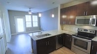 This single-floor unit showcases a well-designed open-concept layout that seamlessly connects the kitchen and living areas. The kitchen serves as the focal point, featuring rich dark-brown cabinetry paired with light granite countertops that extend across both the peninsula and surrounding counters. The peninsula includes an undermount stainless steel sink with a contemporary pull-down faucet, complemented by stylish pendant lighting suspended above. A glass tile backsplash adds a refined touch, while stainless steel appliances, including a dishwasher and range, complete the kitchen's modern aesthetic.

The living area benefits from abundant natural light streaming through multiple windows, creating an airy, open feel. Light-toned hardwood flooring runs throughout, tying the spaces together cohesively. A ceiling fan with integrated lighting enhances airflow and illumination in the living space. The soft blue-gray wall tones provide a neutral, calming backdrop throughout. A door visible from the living area suggests access to a private outdoor space, adding an additional layer of functionality to the unit.