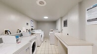This shared laundry facility is a clean, efficiently designed space that accommodates multiple users at once. The room is finished with light-coloured tile flooring and white walls, giving it a bright and sanitary appearance. A row of commercial-grade front-loading washers and top-loading dryers lines one side of the room, all branded and equipped with digital payment terminals, allowing for cashless transactions. A dedicated Coamatic payment station is mounted on the wall, accompanied by a cork bulletin board displaying usage instructions and notices.

A generously sized white folding counter runs along the opposite wall, providing ample surface area for sorting and folding laundry. Ventilation is supported by wall-mounted louvred vents and a ceiling exhaust, ensuring proper air circulation throughout the space. The room is accessed through a sturdy grey metal door marked "Laundry Room." Overhead, flush-mounted ceiling lights provide consistent, even illumination. The layout is practical and uncluttered, with clear signage and instructions posted to guide users. A baseboard heater ensures comfort during cooler months, making this facility a functional and well-maintained amenity.