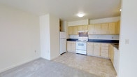 This single-floor residential space offers a practical and straightforward layout designed for comfortable everyday living. The kitchen is a standout feature, outfitted with an abundance of light wood cabinetry that provides generous storage space both above and below the countertops. Dark laminate countertops contrast neatly with the light cabinetry, while white appliances — including a freestanding electric range, a refrigerator, and a range hood — complete the functional cooking area. The kitchen transitions seamlessly into the adjacent living area, separated by a partial wall that defines each zone while maintaining an open, airy feel. The living area is finished with plush, neutral-toned carpeting, contributing to a warm and understated ambiance. The kitchen flooring features light-toned tile, providing a practical and easy-to-maintain surface. Walls throughout are painted in a soft, neutral cream tone, lending the space a clean and bright atmosphere. Recessed ceiling lighting in the kitchen and a flush-mount light fixture ensure the space is well-lit. Overall, the space presents a clean, functional environment well-suited for daily residential use.