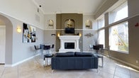This elegantly designed single-floor space immediately impresses with its dramatic double-height ceilings that amplify the sense of openness and grandeur. The central focal point of the living area is a striking white fireplace with a classic mantel, flanked symmetrically by illuminated arched wall niches that display artwork and add architectural character. A mounted flat-screen television above the mantel integrates modern convenience seamlessly into the traditional design. The walls feature a refined two-tone color scheme of warm taupe and soft grey, complemented by crisp crown molding that runs along the upper perimeter, lending a polished, upscale finish. Light pours in through large windows, enhancing the airy ambiance and highlighting the neutral-toned tile flooring that extends throughout the space. The living area is anchored by a sleek black leather sofa and accented with contemporary metallic wall art, creating a sophisticated contrast against the light walls. Bistro-style high-top tables with modern chairs are positioned on either side of the room, offering flexible seating arrangements. The overall design balances classic architectural details with contemporary furnishings, resulting in a stylish, welcoming, and highly functional space.