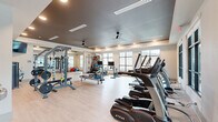 Located at 5697 SW 49th Rd in Ocala, Florida, this fitness center offers a generous, open single-floor layout designed to support a wide range of workout routines. The space features light wood-look flooring throughout, complemented by a two-tone color scheme of soft white walls and a sleek dark gray ceiling detail that lends the room a modern, polished aesthetic. Recessed lighting and ceiling fans are distributed evenly overhead, ensuring both comfort and visibility across the entire floor.

Large multi-pane windows line the exterior walls, flooding the space with abundant natural light and providing views of the surrounding outdoor environment. The cardio zone is well-appointed with a row of True-brand treadmills, ellipticals, stationary bikes, and a rowing machine, each equipped with individual monitors. The strength training area includes a multi-station cable machine, a weight bench with barbell and plate storage, and a full dumbbell rack. Floor space is generous, allowing for stretching and functional training with accessories such as stability balls.

A wall-mounted flat-screen television provides entertainment, while a full-length mirror expands the sense of space and supports form monitoring during workouts. Near the entrance, dual water fountains, a sanitizing station, and a waste receptacle ensure cleanliness and convenience. Posted fitness center rules reflect a well-managed, community-oriented facility.