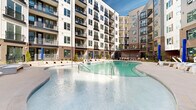 Located at 1133 Harding Place in Charlotte's vibrant 28204 zip code, this outdoor amenity courtyard is a standout feature of a modern multi-story residential community. The space is defined by a generously sized, freeform swimming pool with a smooth concrete surround, offering ample room for lounging and recreation. Rows of wicker-style chaise lounges and umbrellas line the pool deck, providing a resort-like atmosphere ideal for relaxation. The courtyard is enveloped on multiple sides by the building's contemporary façade, which blends brick, white paneling, and large-paned windows with private balconies — creating a sense of enclosure and community while maintaining an open-air feel. Thoughtfully designed landscaping, including ornamental grasses, young trees, and mulched garden beds, adds natural texture and greenery throughout the space. A covered pergola structure at one end of the courtyard provides a shaded gathering area, complementing the open sundeck zones. Black iron fencing with a gated entry defines the perimeter, lending both security and architectural cohesion. The entire space benefits from abundant natural light and open sky views, reinforcing its resort-inspired character in the heart of Charlotte.