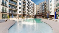 Located at 1133 Harding Place in Charlotte's vibrant 28204 zip code, this outdoor amenity courtyard is a standout feature of a modern multi-story residential community. The space is defined by a generously sized, freeform swimming pool with a smooth concrete surround, offering ample room for lounging and recreation. Rows of wicker-style chaise lounges and umbrellas line the pool deck, providing a resort-like atmosphere ideal for relaxation. The courtyard is enveloped on multiple sides by the building's contemporary façade, which blends brick, white paneling, and large-paned windows with private balconies — creating a sense of enclosure and community while maintaining an open-air feel. Thoughtfully designed landscaping, including ornamental grasses, young trees, and mulched garden beds, adds natural texture and greenery throughout the space. A covered pergola structure at one end of the courtyard provides a shaded gathering area, complementing the open sundeck zones. Black iron fencing with a gated entry defines the perimeter, lending both security and architectural cohesion. The entire space benefits from abundant natural light and open sky views, reinforcing its resort-inspired character in the heart of Charlotte.
