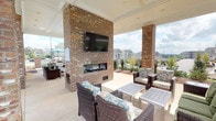 This single-level outdoor amenity area is thoughtfully designed to serve as a multifunctional gathering space, combining covered and open-air sections for year-round enjoyment. The centerpiece of the covered patio is a floor-to-ceiling brick fireplace with a contemporary linear firebox and a wall-mounted flat-screen television above, creating a comfortable lounge atmosphere. Multiple seating arrangements surround this feature, including wicker chairs with cushioned upholstery and low slung side tables, offering ample space for socializing.

Brick columns frame the covered portion of the patio, supporting a white-paneled ceiling while keeping the space open to views of the surrounding landscape. Adjacent to the lounge area, an uncovered patio section extends outward and includes an outdoor kitchen with a built-in grill, counter seating along a railing, and a dining table with chairs shaded by a market umbrella—ideal for outdoor cooking and casual meals.

A spacious concrete pool deck is accessible from the patio, enclosed by a black iron security fence with brick column accents. The pool area features lounge chairs and open sky views. Throughout the space, the consistent use of brick detailing, neutral concrete flooring, and manicured landscaping creates a cohesive, resort-style aesthetic.