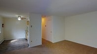 This single-floor residential space offers a straightforward and functional layout designed for comfortable everyday living. The main living area is open and spacious, featuring neutral white walls and carpeted flooring that extend throughout the space, creating a cohesive and understated aesthetic. A ceiling fan is visible in the adjoining area, providing practical ventilation and air circulation.

The kitchen is positioned adjacent to the living space, separated by a partial wall that maintains an open connection between the two areas. The kitchen includes cabinetry and standard appliances, offering a utilitarian setup suited for daily use. The partial wall between the kitchen and living room also provides subtle definition between spaces without fully closing them off.

A hallway extends from the main living area, leading to additional rooms and featuring a white door at its end, suggesting access to a private room or storage area. The hallway is warmly lit, contributing to the overall inviting atmosphere of the interior. The space maintains consistent white walls throughout, lending it a clean and neutral character that provides a versatile backdrop for a variety of furnishing styles. Overall, the space is practical, well-connected, and move-in ready.