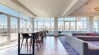 Located at 201 10th Avenue SE in Calgary, the Sky Lounge is an elegantly designed elevated amenity space offering sweeping views of the downtown skyline, including iconic landmarks such as the Calgary Tower. The single-floor layout is generously proportioned, providing a comfortable and versatile environment suited for both social gatherings and relaxed lounging.

Floor-to-ceiling glass windows wrap much of the perimeter, flooding the interior with abundant natural light and framing striking panoramic city views from multiple vantage points. Light-toned hardwood flooring runs throughout, complementing the modern, neutral palette of the space. The lounge is organized into two distinct zones: a lively social area and a more relaxed lounge setting.

The social area features tall bar-height tables paired with upholstered counter stools, ideal for casual conversation or work. A window-side counter with blue bar seating offers additional workspace with unobstructed skyline views. The lounge area is anchored by a large modular sofa, a vibrant area rug, and a dramatic floor-to-ceiling stone-clad fireplace with a wide linear burner, topped by a mounted flat-screen display. Striking circular gold pendant lights add warmth and character overhead.

A kitchenette with cabinetry, a sink, and a full-size refrigerator supports event hosting. Direct access to an outdoor terrace further enhances the appeal of this elevated, design-forward space.