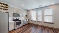 This single-floor space is designed with a clean, modern aesthetic that emphasizes functionality and style throughout. Rich, dark hardwood-style flooring runs continuously across the main living and kitchen areas, providing a warm contrast to the bright white walls and ceilings equipped with recessed lighting.

The kitchen is a standout feature, offering a full suite of stainless steel appliances, including a side-by-side refrigerator with an ice and water dispenser, an electric range, an over-the-range microwave, and a dishwasher. Cabinetry in a soft greige tone lines the walls, complemented by a classic white subway tile backsplash and light countertops. A stainless steel farmhouse-style sink sits beneath a set of windows fitted with white horizontal blinds, allowing ample natural light. A built-in wine rack above the refrigerator adds a thoughtful design detail.

The living area is open and generously proportioned, featuring multiple windows with blinds, a ceiling fan, and recessed lighting that keep the space bright and airy.

The bathroom maintains the contemporary theme with large-format light tile flooring, a glass-enclosed walk-in shower with subway tile surround, modern cabinetry, and clean white countertops.