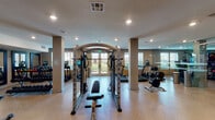 This single-floor fitness center is thoughtfully designed to support a full range of workout routines in a spacious, open environment. The layout features light-toned parquet-style flooring throughout, which contributes to a clean and modern aesthetic. Structural columns are positioned throughout the space without disrupting the flow of the floor plan, and recessed lighting provides consistent, well-distributed illumination across the entire area.

The facility is outfitted with an extensive selection of equipment, including treadmills, a cable machine with an adjustable bench, rowing machines, spin bikes with integrated displays, ellipticals, and a variety of selectorized strength machines. A dedicated dumbbell rack stocked with a range of weights is also present, alongside stability balls and additional free weight accessories.

Large floor-to-ceiling mirrors line multiple walls, enhancing the sense of space and allowing users to monitor their form during exercise. A full wall of glass-paneled doors runs along one side, adding an elegant architectural detail and connecting the interior to exterior views featuring palm trees and outdoor surroundings. Additional windows allow natural light to pour in, creating a bright and energizing atmosphere.

Dual water fountains are conveniently mounted along one wall, and the space includes clearly marked exit access points. The overall design balances functionality with a polished, upscale aesthetic suited for serious fitness use.