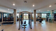 This single-floor fitness center is thoughtfully designed to support a full range of workout routines in a spacious, open environment. The layout features light-toned parquet-style flooring throughout, which contributes to a clean and modern aesthetic. Structural columns are positioned throughout the space without disrupting the flow of the floor plan, and recessed lighting provides consistent, well-distributed illumination across the entire area.

The facility is outfitted with an extensive selection of equipment, including treadmills, a cable machine with an adjustable bench, rowing machines, spin bikes with integrated displays, ellipticals, and a variety of selectorized strength machines. A dedicated dumbbell rack stocked with a range of weights is also present, alongside stability balls and additional free weight accessories.

Large floor-to-ceiling mirrors line multiple walls, enhancing the sense of space and allowing users to monitor their form during exercise. A full wall of glass-paneled doors runs along one side, adding an elegant architectural detail and connecting the interior to exterior views featuring palm trees and outdoor surroundings. Additional windows allow natural light to pour in, creating a bright and energizing atmosphere.

Dual water fountains are conveniently mounted along one wall, and the space includes clearly marked exit access points. The overall design balances functionality with a polished, upscale aesthetic suited for serious fitness use.