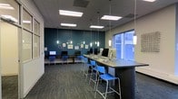 This single-floor space functions as a well-equipped computer lab designed to support both individual focus and collaborative work. The room is anchored by a bold teal feature wall adorned with framed artwork and circular decorative elements, adding visual interest and character to the functional environment. Two distinct work zones define the layout: a row of traditional desktop computer workstations along one wall, paired with standard seating, and a long bar-height counter running through the center of the room, lined with vibrant blue chairs that encourage a more casual, collaborative style of working. Desktop computers and monitors are distributed throughout both areas, ensuring ample access to technology. Overhead, a combination of recessed panel lighting and suspended pendant lights provides layered illumination across the space. A large window along one wall allows natural light to filter in, brightening the room and offering a connection to the exterior. The patterned carpet flooring and drop-tile ceiling contribute to a clean, organized aesthetic. Overall, the space strikes a balance between a professional and approachable atmosphere, well-suited for learning, research, or tech-focused collaboration.