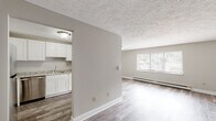 This single-floor unit offers a clean, functional layout with a neutral color palette and wood-look vinyl flooring that flows throughout the main living areas. The open living room is bathed in natural light through a large picture window, complemented by a baseboard heating system and a wall-mounted air conditioning unit, ensuring year-round comfort. A textured ceiling adds character to the space. The adjacent kitchen is efficiently designed in an L-shaped configuration, featuring crisp white shaker-style cabinetry, light granite countertops, a stainless steel sink, and a full suite of stainless steel appliances including a refrigerator, electric range, over-the-range microwave, and dishwasher. The bathroom is outfitted with a full soaking tub and shower combination, a toilet, built-in shelving nook, and matching wood-look flooring for a cohesive aesthetic. The bedroom is finished with soft wall-to-wall carpeting and includes a generously sized reach-in closet with a full-width hanging rod, providing ample storage. The overall design is practical and well-maintained, making this a straightforward and comfortable single-level living space.