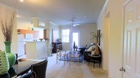 This single-floor residence offers an open-concept layout that seamlessly connects the main living areas, creating a cohesive and functional living environment. The living room serves as the central gathering space, featuring plush wall-to-wall carpeting, crown molding, and a ceiling fan that enhances air circulation throughout the room. Natural light flows in through multiple windows, including a bright glass door that adds to the airy atmosphere of the space. The living room is anchored by a comfortable seating arrangement complemented by a glass-top coffee table and tasteful decorative accents. Adjacent to the living area, a defined dining space provides room for casual entertaining, with bar-style seating connecting it to the kitchen. The kitchen features a raised breakfast bar that doubles as a partition, offering both a visual separation and a convenient casual dining option. The neutral color palette of warm beige tones throughout the walls and flooring creates a cohesive and inviting ambiance. Crown molding details add a refined architectural touch, elevating the overall aesthetic of the space. The layout is thoughtfully designed to maximize both comfort and functionality, making it ideal for everyday living and casual entertaining.