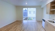 This single-floor space offers a clean, modern interior defined by light-toned hardwood flooring that runs throughout the open living area, lending warmth and continuity to the layout. The room is generously proportioned, providing a comfortable sense of openness well-suited to a variety of furniture arrangements.

A large sliding glass door serves as the focal point of the main living area, fitted with horizontal blinds for privacy and light control. The door allows abundant natural light to flood the interior while offering a direct view of the surrounding neighborhood, including adjacent rooftops and greenery. This feature also provides convenient outdoor access.

The walls are finished in a soft, neutral cream tone that complements the light flooring and contributes to the room's airy, welcoming atmosphere. Recessed ceiling lighting provides supplemental illumination, and clean white baseboards add a crisp, finished look throughout. A partial view of what appears to be white appliances at the room's edge suggests an adjacent kitchen area. Overall, the space presents a straightforward, well-maintained interior with practical features and good natural light.