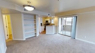 This single-floor residence offers a well-designed, open-concept layout that connects the living area and kitchen in a cohesive, functional arrangement. The living room is finished with soft, neutral wall-to-wall carpeting and features a set of built-in shelving units that provide both storage and display space, adding a refined architectural detail to the room. A contemporary flush-mount pendant light fixture illuminates the space, complementing the warm yellow-toned walls throughout.

The kitchen is positioned at the heart of the unit, centered around a peninsula with a granite-style countertop that serves as both a prep surface and casual dining area. White cabinetry lines the walls, and stainless steel appliances — including a microwave and range — add a modern, polished finish. Wood-style flooring in the kitchen contrasts nicely with the carpeted living area, clearly delineating the two zones.

Large sliding glass doors flood the living space with natural light and provide direct access to a private balcony, offering an outdoor retreat with views of the surrounding exterior. The overall ambiance is clean, bright, and move-in ready, with a neutral palette that lends itself well to a variety of interior styles.