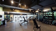 Located at 26 E Pearson St in Chicago's Streeterville neighborhood, this fitness center offers a well-equipped, single-floor workout facility with a sleek, modern aesthetic. The space features an exposed dark ceiling with industrial-style track lighting and ductwork, creating a focused and energetic atmosphere throughout.

The main workout area is outfitted with rubber tile flooring designed for both safety and durability. A dedicated cardio zone lines the perimeter near large floor-to-ceiling windows, housing treadmills, stationary bikes, elliptical trainers, and a rowing machine — all positioned to take full advantage of sweeping views of the Chicago skyline. Natural light streams in generously, making cardio sessions feel open and invigorating.

The strength training section features a multi-station cable machine, adjustable benches, and a full rack of dumbbells organized along a mint-green accent wall, adding a vibrant contrast to the otherwise dark color palette. A water fountain station is conveniently integrated into the same area for easy access during workouts.

A glass-enclosed room provides a separate, dedicated space for additional training or stretching. The facility also includes wall-mounted mirrors to maximize the sense of space and support proper form. The overall design balances industrial urban style with functional fitness infrastructure, making it a comprehensive workout destination in the heart of Chicago.