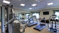 This fitness room offers a thoughtfully arranged workout space designed to accommodate a variety of exercise routines. The room is outfitted with multiple treadmills, an elliptical machine, a recumbent-style strength training unit, and additional cardio equipment, providing options for both cardiovascular and resistance-based workouts. A large wall mirror spans one side of the room, visually expanding the space and allowing users to monitor their form during exercise. Exercise balls are also available, adding versatility to workout options. The drop ceiling features evenly spaced recessed lighting panels that provide consistent, bright illumination throughout. Natural light enters through windows dressed with elegant green valances, contributing a welcoming ambiance to the functional space. A wall-mounted flat-screen television is positioned for easy viewing during workouts. The light-colored walls and neutral flooring create a clean, open atmosphere. The overall design balances practicality with comfort, making it a well-rounded facility suited for regular fitness use.