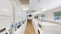 Located at 420 N Dean Rd in Auburn, Alabama, this commercial laundry facility offers a clean, efficiently organized space designed for high-volume use. The single-floor layout is finished in a bright, neutral palette of whites and soft grays, creating a welcoming and hygienic atmosphere throughout.

The laundry room is the centerpiece of the facility, lined with a row of commercial-grade front-loading washers and top-loading machines along one wall, complemented by a bank of stacked dryer units. A central folding counter runs the length of the room, providing ample workspace for sorting and folding. The light wood-look flooring adds warmth to the otherwise crisp, functional interior.

Natural light flows in through large windows that look out onto the surrounding greenery, keeping the space airy and pleasant. Overhead, a series of flush-mount and oval ceiling light fixtures ensure consistent, bright illumination throughout the day and evening. A ceiling-mounted HVAC cassette unit maintains comfortable climate control year-round.

The facility also includes a kitchen area and is equipped with a wall-mounted television, a vending machine, and clearly posted informational signage. An emergency exit door provides convenient access. The overall design prioritizes ease of use, cleanliness, and comfort, making this a practical and well-appointed laundry facility.