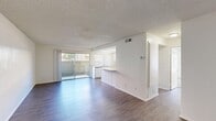 Located at 2098 West Acacia Avenue in Hemet, California, this single-story one-bedroom unit offers a thoughtfully arranged floor plan that maximizes its compact footprint. The open-concept living room greets you with wood-style flooring, crisp white walls, and a textured ceiling. A sliding glass door floods the space with natural light and provides direct access to a private patio, offering pleasant outdoor views. The living area flows seamlessly into the kitchen, which is separated by a peninsula counter with granite-style laminate surfaces — ideal for casual dining or additional prep space. The kitchen is well-appointed with white cabinetry reaching the ceiling, a gas range, double sink, dishwasher, and a bright drop-panel ceiling that enhances visibility throughout. Two hallways connect the living spaces to the private areas of the unit. The generously sized primary bedroom features plush carpeting, a large sliding-door closet, and a window that brings in ample natural light. It also provides direct access to the bathroom, which includes a vanity with cabinetry and a well-lit mirror. Throughout the unit, a neutral color palette and clean finishes create a bright, functional, and move-in-ready atmosphere.