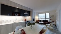 This single-floor residence showcases a thoughtfully designed open-concept layout that seamlessly connects the kitchen, dining, and living areas into one cohesive living space. The kitchen is outfitted with two-tone cabinetry — dark upper cabinets contrasting with light lower units — paired with a stainless steel microwave and range, an undermount sink, and light-colored countertops that extend into a generous kitchen island doubling as a dining surface. Recessed lighting illuminates the kitchen area, complementing the under-cabinet lighting that brightens the workspace. The dining area is anchored by the marble-finish island with seating for multiple guests. The living area beyond features a deep burgundy sofa and coordinating armchair arranged around a glass-top coffee table, creating an inviting lounge setting. A large picture window floods the living space with natural light and offers open outward views, enhancing the airy feel of the interior. Neutral wall tones and white ceilings unify the space, while carefully chosen accents — including red kitchen accessories and blue decorative pieces — add personality. The overall ambiance is clean, contemporary, and functional.