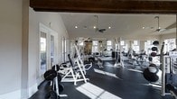 This fitness center offers a comprehensive and thoughtfully designed workout environment suited for a wide range of training needs. The space is anchored by soaring vaulted ceilings with exposed wooden beams, lending a sense of openness and volume to the room. Recessed lighting combined with large windows along multiple walls floods the interior with abundant natural light, creating an inviting and energizing atmosphere. A ceiling fan provides additional airflow throughout the space.

The workout floor is finished with durable dark rubber flooring, providing excellent traction and cushioning for strength and conditioning activities. A full array of white-framed strength training machines and cable systems are arranged throughout the space, complemented by free weights including barbells and weight plates. Adjustable benches and incline racks offer versatility for a variety of exercise routines. Full-length mirrors along one wall enhance the sense of space while allowing users to monitor form during workouts.

Natural light streams through the windows, casting bright patterns across the floor and highlighting the clean, well-maintained equipment. Glass-paned entry doors open to an outdoor area, further enhancing the connection to the surrounding environment. The overall design balances functionality with an open, airy aesthetic, making this fitness center both practical and pleasant to use.