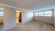 Located at 166 North McCarrons Boulevard in Roseville, MN, this 2-bedroom, 1-bathroom unit offers a straightforward and functional single-floor layout. The spacious primary bedroom features wall-to-wall carpeting, neutral toned walls with a textured finish, and a wide multi-pane window that allows ample natural light to fill the room. A baseboard heating system runs along the wall for year-round comfort. The second bedroom is equally well-appointed with soft carpeting, a built-in wood wardrobe with overhead storage, and a window overlooking greenery outdoors. The open living area features the same warm carpeting and textured walls, with ceiling-mounted lighting and doorways connecting to the kitchen and bedrooms. The kitchen is equipped with slate-gray cabinetry, light stone-toned countertops, a double-basin sink, and a full-size refrigerator, all complemented by wood-look flooring and a window providing natural light. The bathroom is tiled throughout with natural stone-patterned wall tiles surrounding a full shower-tub combination, a pedestal sink, and a glass block window that brings in diffused light. A hallway connects the key areas of the unit, providing a cohesive and easy-to-navigate flow throughout the space.