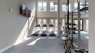 This well-appointed fitness center is designed to support a full spectrum of workout routines within a bright, open environment. The space is defined by its striking double-height ceilings, which lend an airy, expansive quality throughout. Large floor-to-ceiling windows and glass doors line the perimeter, flooding the interior with natural light and providing views of the surrounding outdoor greenery.

The layout is thoughtfully organized into distinct training zones. The cardio area features a row of treadmills positioned directly in front of the windowed wall, alongside elliptical machines and upright bikes. A dedicated strength training section includes a full dumbbell rack with a range of weights, adjustable benches, and a cable machine, all set against a wall of mirrors with wall-mounted televisions overhead.

A functional training zone offers a multi-use rig with TRX suspension straps, a heavy punching bag, and open floor space equipped with exercise mats and balance trainers. Additional wall-mounted screens are positioned throughout the space, ensuring visibility from all workout areas.

The neutral color palette of warm grey tones, white columns, and carpet tile flooring contributes to a clean, professional atmosphere that is both motivating and comfortable.