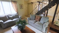 This two-story residence offers a thoughtfully designed interior that balances functionality with a warm, inviting atmosphere. The living room serves as the heart of the main level, characterized by vaulted ceilings that lend a sense of openness and volume to the space. A large window dressed with floral curtains allows generous natural light to fill the room, creating a bright and airy environment. The living area comfortably accommodates seating arranged around a wooden coffee table, complemented by an abundance of indoor plants that add a natural, organic touch to the décor. A pendant light fixture overhead provides ambient illumination, enhancing the room's welcoming character. Adjacent to the living space, a defined entryway features a front door with a decorative rug and framed wall art nearby, establishing a clear transition point into the home. A staircase with a wrought iron railing is visible from the living area, providing access to the second floor. The carpeted flooring throughout the main living space adds warmth and comfort underfoot, while the neutral wall tones create a versatile backdrop suitable for a variety of interior styles.
