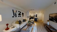 This single-floor residence offers a thoughtfully designed open-concept layout that seamlessly connects the living, dining, and kitchen areas into one cohesive and functional space. The living area is anchored by a clean-lined white sofa accented with mustard and charcoal throw pillows, complemented by a round wooden coffee table and a geometric patterned area rug in warm tones of gold, white, and gray. Bold abstract wall art adds a contemporary artistic touch to the space. Recessed lighting and track lighting illuminate the interior, enhancing the bright and airy atmosphere created by the light-toned walls and smooth ceilings. The kitchen features white upper cabinetry, a stainless steel refrigerator, and dark lower cabinetry with ample counter space, creating a sleek two-tone aesthetic. A kitchen island with bar seating provides a casual dining option, finished with a pendant light overhead. Warm-toned hardwood flooring flows throughout the entire space, tying all areas together cohesively. The hallway leads to additional spaces beyond, maintaining a clean, modern aesthetic. Overall, the space exudes a contemporary and well-curated ambiance suited for comfortable, stylish living.