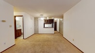 This single-floor unit on W Hampton Avenue in Milwaukee, Wisconsin offers a straightforward, functional layout spread across several well-defined spaces. The living area is generously proportioned, featuring neutral wall-to-wall carpeting, white walls with wood baseboards, and a ceiling fan with light fixture overhead. A wall of louvered bifold closet doors provides notable built-in storage within the living space. The kitchen is positioned centrally with a pass-through counter connecting it to the main living area. Dark walnut-toned cabinetry lines the upper walls, complemented by a white gas range, wall-mounted ventilation fan, and ample counter space. The bedroom areas each feature carpeted floors, natural window light, baseboard heating, and louvered closet doors with shelving and hanging rods. One bedroom includes a walk-in closet accessible through a dark wood door. The full bathroom is appointed with a dark wood vanity, white countertop, large mirror, mosaic tile flooring, and a full tub with white tile surround. Throughout the unit, the design maintains a clean, understated aesthetic with consistent finishes.