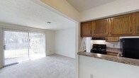This single-floor residence is designed with a straightforward, functional layout that maximizes the use of its interior space. The home features soft, neutral-toned walls throughout, complemented by light carpet flooring that extends across the main living areas and hallway, creating a cohesive and understated aesthetic.

The living area is open and bright, benefiting significantly from a full-height sliding glass door fitted with vertical blinds, which allows natural light to fill the space and provides a view of the greenery outside. The textured ceiling adds subtle visual interest overhead, while a ceiling vent ensures comfortable climate control.

Adjacent to the living area, the kitchen is efficiently arranged with warm-toned wooden cabinetry offering ample storage. A black electric range sits beneath an overhead exhaust hood, and a peninsula with a laminate countertop separates the kitchen from the living space, creating a natural divide while maintaining an open feel.

A hallway connects the main living areas to the remaining rooms, featuring a paneled door—likely a closet—adding practical storage. The overall ambiance of the home is clean, neutral, and move-in ready, with a calm and uncluttered atmosphere throughout.