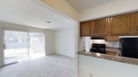 This single-floor residence is designed with a straightforward, functional layout that maximizes the use of its interior space. The home features soft, neutral-toned walls throughout, complemented by light carpet flooring that extends across the main living areas and hallway, creating a cohesive and understated aesthetic.

The living area is open and bright, benefiting significantly from a full-height sliding glass door fitted with vertical blinds, which allows natural light to fill the space and provides a view of the greenery outside. The textured ceiling adds subtle visual interest overhead, while a ceiling vent ensures comfortable climate control.

Adjacent to the living area, the kitchen is efficiently arranged with warm-toned wooden cabinetry offering ample storage. A black electric range sits beneath an overhead exhaust hood, and a peninsula with a laminate countertop separates the kitchen from the living space, creating a natural divide while maintaining an open feel.

A hallway connects the main living areas to the remaining rooms, featuring a paneled door—likely a closet—adding practical storage. The overall ambiance of the home is clean, neutral, and move-in ready, with a calm and uncluttered atmosphere throughout.