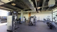 This fitness facility offers a spacious, open-plan workout environment designed to accommodate a comprehensive range of exercise needs. The space features an exposed industrial ceiling with visible ductwork and structural beams finished in dark tones, lending it a modern, urban aesthetic. Pendant lighting fixtures are distributed throughout the ceiling, providing ample illumination across the workout floor.

The flooring consists of dark rubber matting, a practical and durable surface well-suited for heavy equipment and high-traffic use. A robust collection of strength training machines is arranged throughout the main floor, including cable machines, multi-functional weight stations, and resistance equipment suitable for targeting various muscle groups. A medicine ball rack and additional cardio equipment are visible along the perimeter walls.

Toward the rear of the space, a secondary area is partially enclosed, housing additional equipment and storage, with lighter wall tones that contrast the darker main floor aesthetic. The overall layout is thoughtfully organized to allow users to move freely between stations. The combination of industrial design elements, quality equipment, and practical flooring makes this a functional and well-appointed fitness space suited for a variety of training styles and fitness levels.