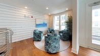 This thoughtfully designed single-floor space exudes a clean, coastal-inspired aesthetic that balances comfort with contemporary style. The interior is anchored by rich, wide-plank hardwood flooring that flows throughout, lending warmth to the bright, white surroundings.

A standout feature is the horizontal shiplap accent wall, which serves as a gallery-style backdrop for curated abstract artwork rendered in calming shades of blue and white — a cohesive artistic thread that runs through the space. Recessed lighting overhead complements the abundant natural light that pours in through large, curtained windows, offering views of palm trees and neighboring architecture.

The lounge area is centered around a circular area rug, where a set of plush, blue mosaic-patterned swivel chairs are arranged invitingly around a low coffee table — ideal for conversation and gathering. A tall, lush potted palm adds a natural, organic element to the corner, enhancing the airy, resort-like atmosphere.

A secondary open area provides additional flexible space, featuring sleek stainless steel cable railings along a mezzanine-style overlook, adding a modern architectural dimension. White paneled doors and clean baseboards throughout contribute to the space's polished, well-maintained character. Overall, the space presents a welcoming, upscale communal environment with a distinctly coastal sensibility.