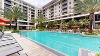 Located at 1 Main Street in Miramar, Florida, this expansive outdoor amenity courtyard is designed to deliver a resort-like experience within a modern multi-story residential community. The centerpiece is a generously sized swimming pool with crystal-clear turquoise water, framed by wide-format stone pavers and lined with sleek lounge chairs and vibrant orange umbrellas for sun-soaking comfort. Adjacent to the pool, a dedicated hot tub is nestled among lush tropical plantings, offering a more intimate relaxation option. The landscaping throughout the courtyard is a standout feature, showcasing an impressive collection of tall royal palms, monstera, philodendron, bromeliads, and tropical ground cover that create a rich, verdant atmosphere. A covered pergola area strung with bistro lighting provides a shaded outdoor dining and social gathering space. An outdoor shower station, finished with sleek wood-slat paneling and pebble tile flooring, adds a practical poolside amenity. Covered walkways with warm wood-paneled ceilings connect various areas of the courtyard, offering shade and architectural continuity. The surrounding building facades incorporate living green walls, further enhancing the tropical aesthetic. The overall ambiance is vibrant, open, and resort-inspired, ideal for outdoor leisure and community living.