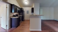This single-floor residence at Rosemeade Apartments in Southbridge, Massachusetts, offers a thoughtfully designed layout with consistent finishes throughout. Rich hardwood flooring flows across the living areas, complemented by vaulted ceilings that lend an open, airy quality to the space.

The kitchen is a functional centerpiece, outfitted with dark espresso cabinetry, granite-style countertops, and stainless steel appliances including a range, microwave, and refrigerator. A peninsula counter provides additional prep space and serves as a natural divider between the kitchen and the adjoining living area. The electrical panel is conveniently accessible within the kitchen.

The bedroom features vaulted ceilings, a ceiling fan with light fixture, a baseboard heating unit, and a window that frames views of surrounding greenery, allowing natural light to fill the room. A thermostat and ventilation are also present.

The bathroom is finished with dark cabinetry, granite-style countertops, an undermount sink, a large mirror, and hardwood-style flooring. In-unit laundry is also available, adding to the overall convenience of the space. Baseboard heating is present throughout, ensuring year-round comfort.
